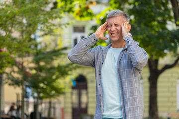 Displeased exhausted Caucasian middle-aged man rubbing temples to cure headache problem suffering from tension migraine stress. Mature male guy tourist grimacing, pain standing on city urban street.
