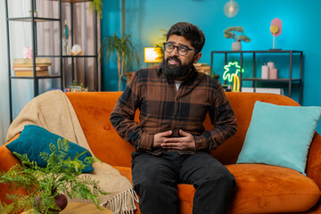 Fototapeta premium Indian young man sitting on sofa feeling sudden strong abdominal stomach ache, gastritis problem. Upset Arabian guy having symptom poisoning diarrhea indigestion peptic ulcer pancreatitis at home room