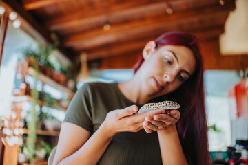 Young woman carefully handles a lizard in her home, surrounded by plants and warm wooden interiors,...