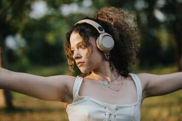 A young woman performs a modern dance in a sunlit park, wearing headphones and expressing joy and...
