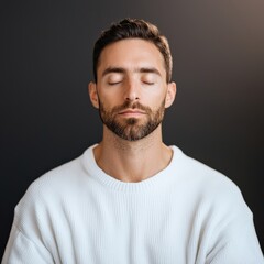 Obraz premium A serene man with a beard and closed eyes, wearing a white sweater, exudes calmness against a dark background.