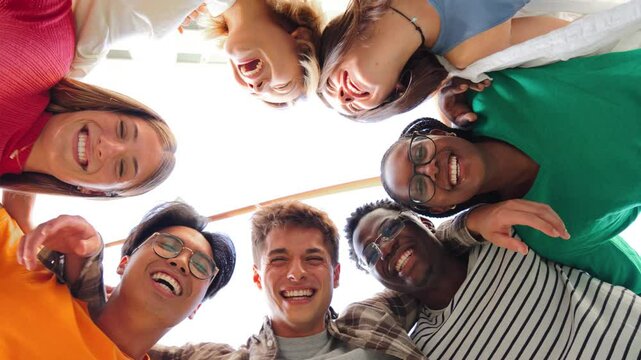 Low angle view of a diverse group of young people forming a circle, smiling and looking down at the camera, symbolizing friendship, teamwork, unity, and happiness in a cheerful and inclusive