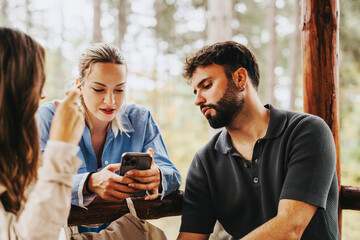 A group of young friends gather outdoors, engaging with a smart phone. They are sitting close, sharing a moment of social interaction and connectivity in a natural setting.