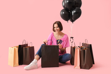 Beautiful young woman with credit cards, shopping bags and air balloons sitting on pink background....
