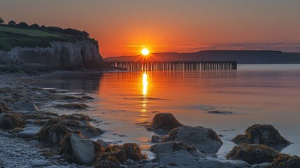 Obraz premium Serene sunset over a calm shoreline with a pier reflecting in the water.