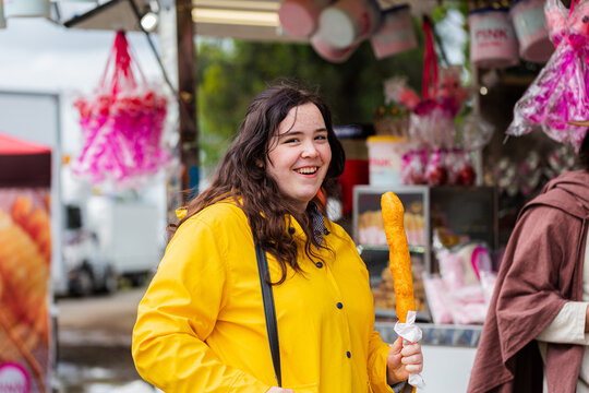 Smiling young woman in yellow rain jacket eating fair food at local agricultural show