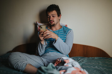 Fototapeta premium A young man in casual pajamas sits on his bed with a smart phone in hand, appearing thoughtful. The setting suggests a relaxed morning or evening routine.