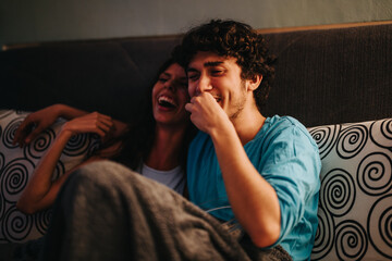 Fototapeta premium A joyful young couple shares a laugh while sitting on a cozy sofa covered in patterned pillows. Their close bond and happiness are evident, creating a warm and inviting atmosphere.