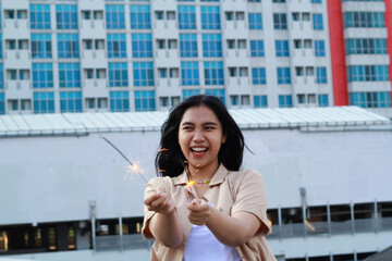 Fototapeta premium carefree asian young woman holding sparkler celebrate new year eve with laugh and dance in rooftop apartment outdoor with city building background