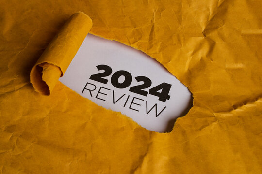 A yellow piece of paper with a hole torn in it. Through the hole, a white piece of paper is visible with the text "2024 REVIEW" written in black. This represents the concept of reviewing the year 2024