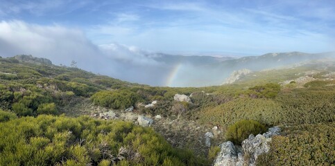 Panoramic view of a lush green mountain landscape with a faint rainbow and misty clouds under a blue sky