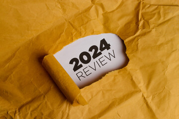 A yellow piece of paper with a hole torn in it. Through the hole, a white piece of paper is visible with the text "2024 REVIEW" written in black. This represents the concept of reviewing the year 2024