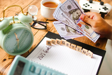 Fototapeta premium 2025 Budgeting - A person planning their budget with money, alarm clock, tea, gamepad, and notebook, emphasizing the importance of financial planning and goal-setting for the future