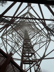 Fototapeta premium A striking upward view of a tall metal communication tower, framed by foggy skies and intricate lattice beams, emphasizing its height and structure.