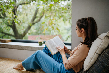 Hygge moment for beautiful woman sitting by a large window with a book. Woman is enjoying time for...