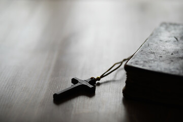 Close-up of a Black Cross Pendant on a Wooden Surface with an Old Leather-bound Book in Soft Focus...
