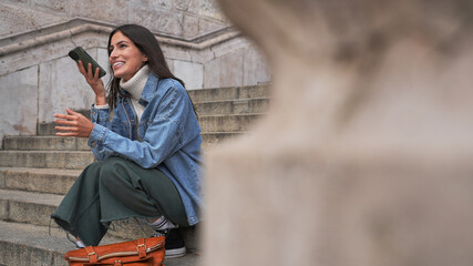 A woman sits on outdoor steps, dropping a voice message on her phone. Travel and technology concept