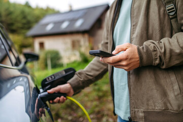 Man charging electric car in front of his house, plugging the charger into the charging port. House...