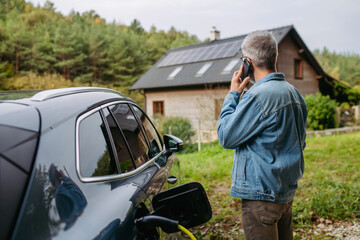 Man charging electric car in front of his house while making phone call. House with solar panel...