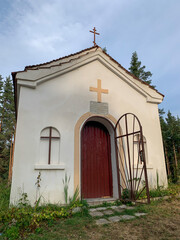 A quaint, small chapel in Bulgaria stands with a simple yet charming facade, featuring a red wooden door, a cross above the entrance, and a partially open wrought iron gate.