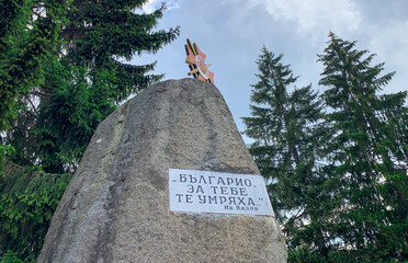 A memorial plate in Batak, Bulgaria, is mounted on a large rock with a plaque (memorial plate), surrounded by tall evergreen trees and topped with a decorative emblem.