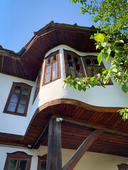 The facade of the old Bulgarian traditional house showcases charming wooden eaves, white plaster walls, and wooden-framed windows, partially hidden by lush green foliage