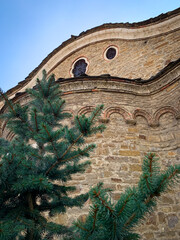 The weathered stone facade of the Saint Nikola church in Dryanovo, Bulgaria, with its distinctive arched windows and a small pine tree in the foreground, evokes a sense of timeless beauty