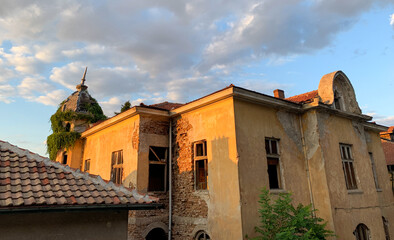 The sun casts a warm glow on an old, weathered mansion with a crumbling facade and a distinctive tower.