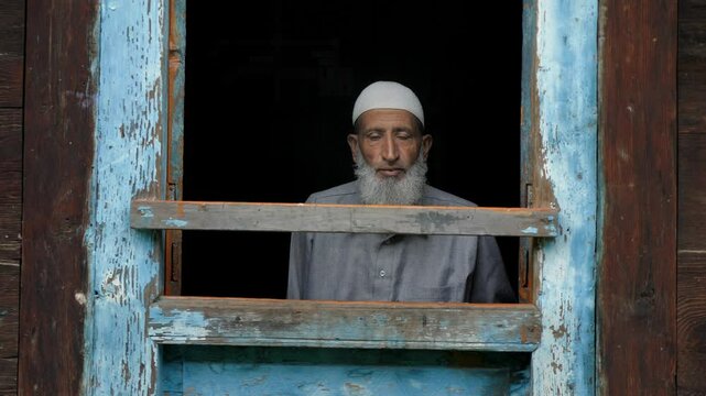 An aged or elderly Muslim Indian Kashmiri man in ethnic attire sitting indoors, looking outside from the frame of the window of an old village wooden house in a Himalayan mountainous region of Kashmir