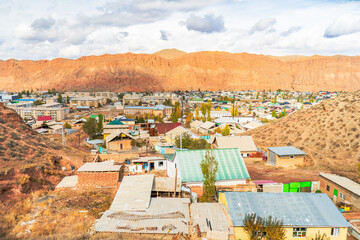 A vibrant town nestled against a backdrop of striking red rock mountains. The scene showcases a mix of modest homes, colorful rooftops, and natural beauty under a partly cloudy sky.