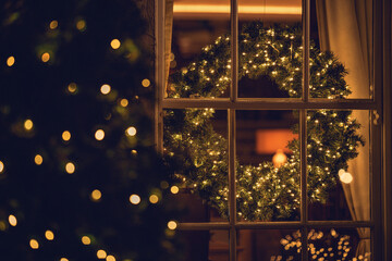 Close-up view through a window of an illuminated Christmas wreath in a living room with an illuminated Christmas tree outdoors