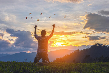 Young male kneeling down with hands open palm up praying to God on the mountain sunset background.