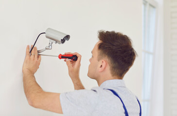 Young technician man in uniform installing surveillance CCTV camera on the wall indoors. Male...