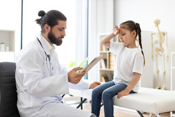 Obraz premium Caucasian male doctor checks little girl with headache at medical clinic. Young patient shows discomfort while doctor reviews information on tablet in professional environment.