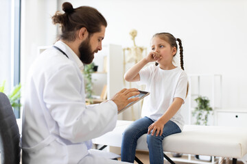 Obraz premium Caucasian male doctor consults young girl sitting on clinic bed. Girl appears to be coughing, indicating a medical examination in progress. Doctor uses tablet for digital notes with patient.