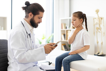 Obraz premium Caucasian male doctor with beard examines Caucasian young girl complaining of sore throat in medical office. Doctor checks symptoms using digital device while girl holds throat.