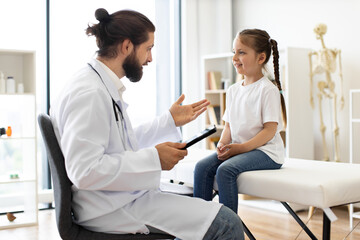 Obraz premium Male doctor talks with young white girl sitting on medical couch, recording details in tablet. Scene in bright examination room with relaxed and comfortable atmosphere.