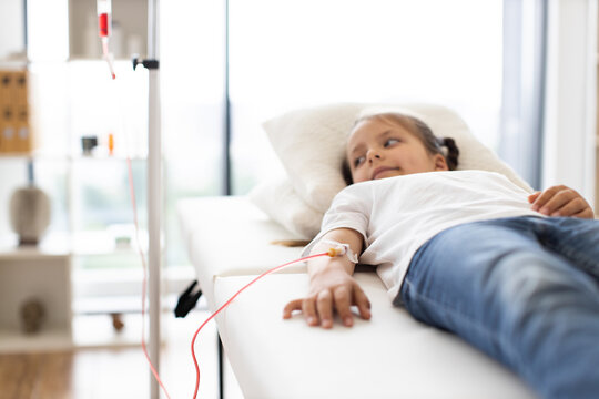 Young Caucasian girl lies on medical couch receiving blood transfusion. She looks calm and comfortable in hospital room. Child wears casual clothing while connected to IV drip, highlighting healthcare - Powered by Adobe