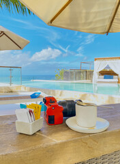 Camera, coffee sachets and cup of coffee drinks on the wooden table at luxury resort infinity pool...