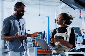 Engineer in car service uses laptop to calculate invoice after fixing broken automobile brakes. Adept garage expert uses device to inform client of final costs after mending vehicle