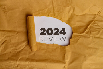 A yellow piece of paper with a hole torn in it. Through the hole, a white piece of paper is visible with the text "2024 REVIEW" written in black. This represents the concept of reviewing the year 2024