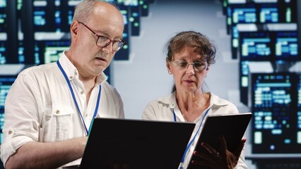 Dilligent manager and assistant looking around data center, using laptop and tablet to crosscheck disaster recovery plan and assess high tech workspace units in need of replacement, preventing risks