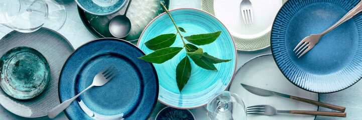 Modern tableware banner, overhead flat lay panoramic shot. A table setting with plates, cutlery, and glasses, shot from the top with green leaves