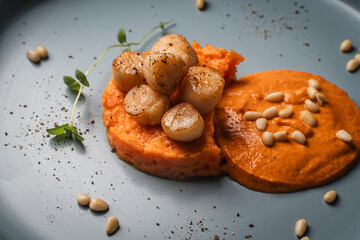 Scallops with carrot puree with Romesco sauce, fesalis and pine nuts on a blue plate on a light...