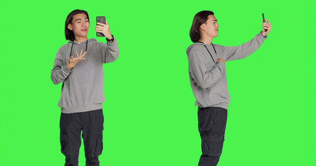 Asian man on videocall connection with friends, using smartphone to call people using phone webcam against greenscreen. Male model talking to someone on videoconference network.