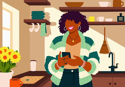 Cheerful young woman standing and using phone in kitchen