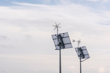 Na zdjęciu widać tył dwóch instalacji wiatrowo-słonecznych. Panele słoneczne zamontowane na masztach nachylone są ku dołowi. Nad nimi znajdują się turbiny wiatrowe. W tle jasne niebo z lekkimi chmuram © Marcin
