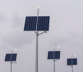 Na zdjęciu widać 4 zestawy złożone z 4 paneli słonecznych i 4 turbin wiatrowych. Panele są granatowe, prostokątne, zamontowane na wysokich masztach, a nad nimi znajdują się białe wiatraki. W tle szare