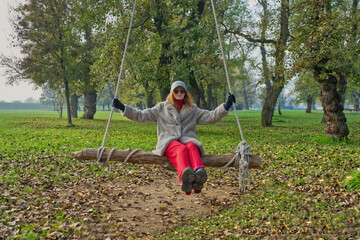 A young girl is swinging on a rustic wooden swing in a park. The swing is made from a log and suspended by thick ropes