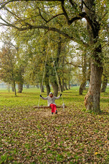 A girl is sitting on a wooden swing suspended from a tree in a park. The ground is covered with fallen leaves, and the surrounding trees have autumn foliage.